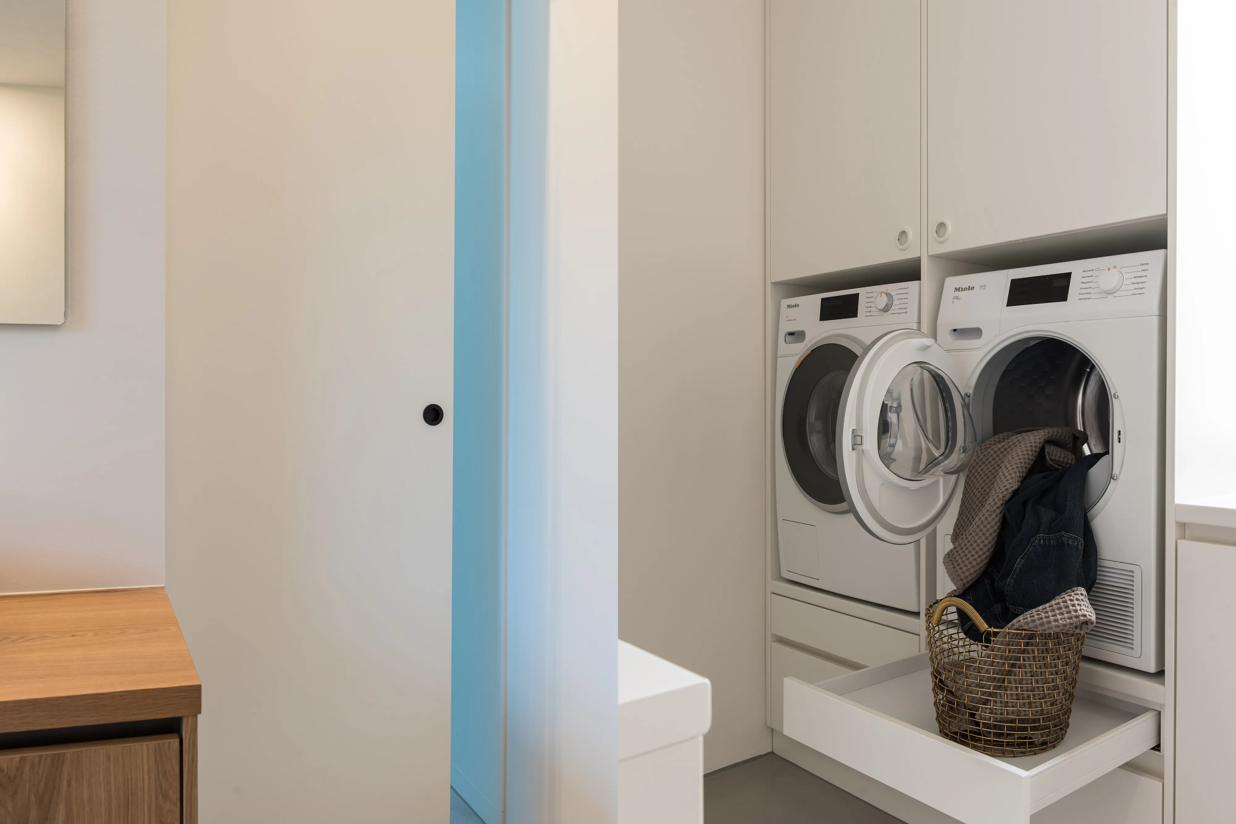 Hauswirtschaftsraum mit erhöht eingebauter Waschmaschine und Wäschetrockner in weißen Einbauschränken sowie ausziehbarem Wäschekorb