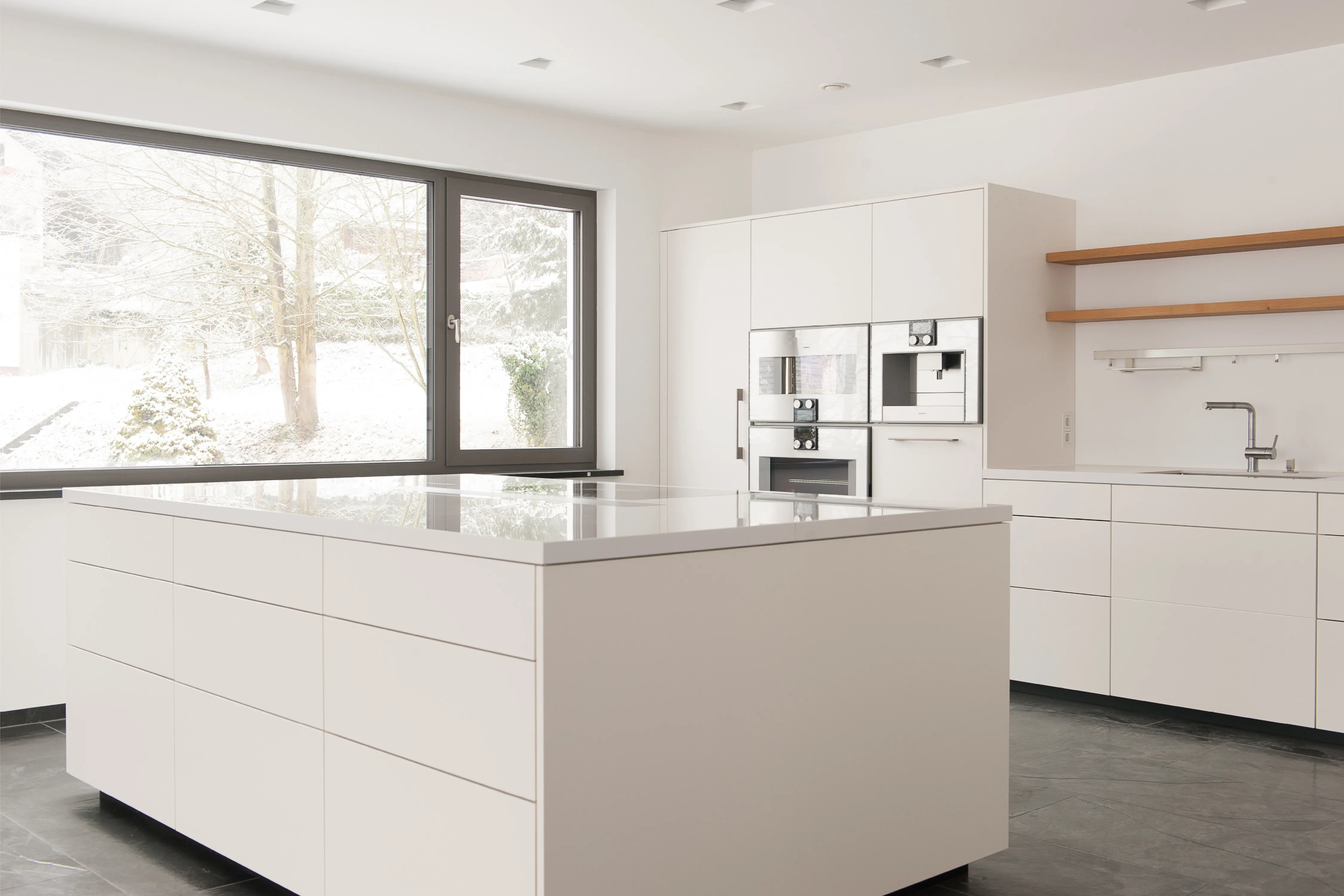 Großzügige Kücheninsel mit weißer Arbeitsplatte vor einer Einbauküche mit Holzwandregalen und Blick ins Verschneite