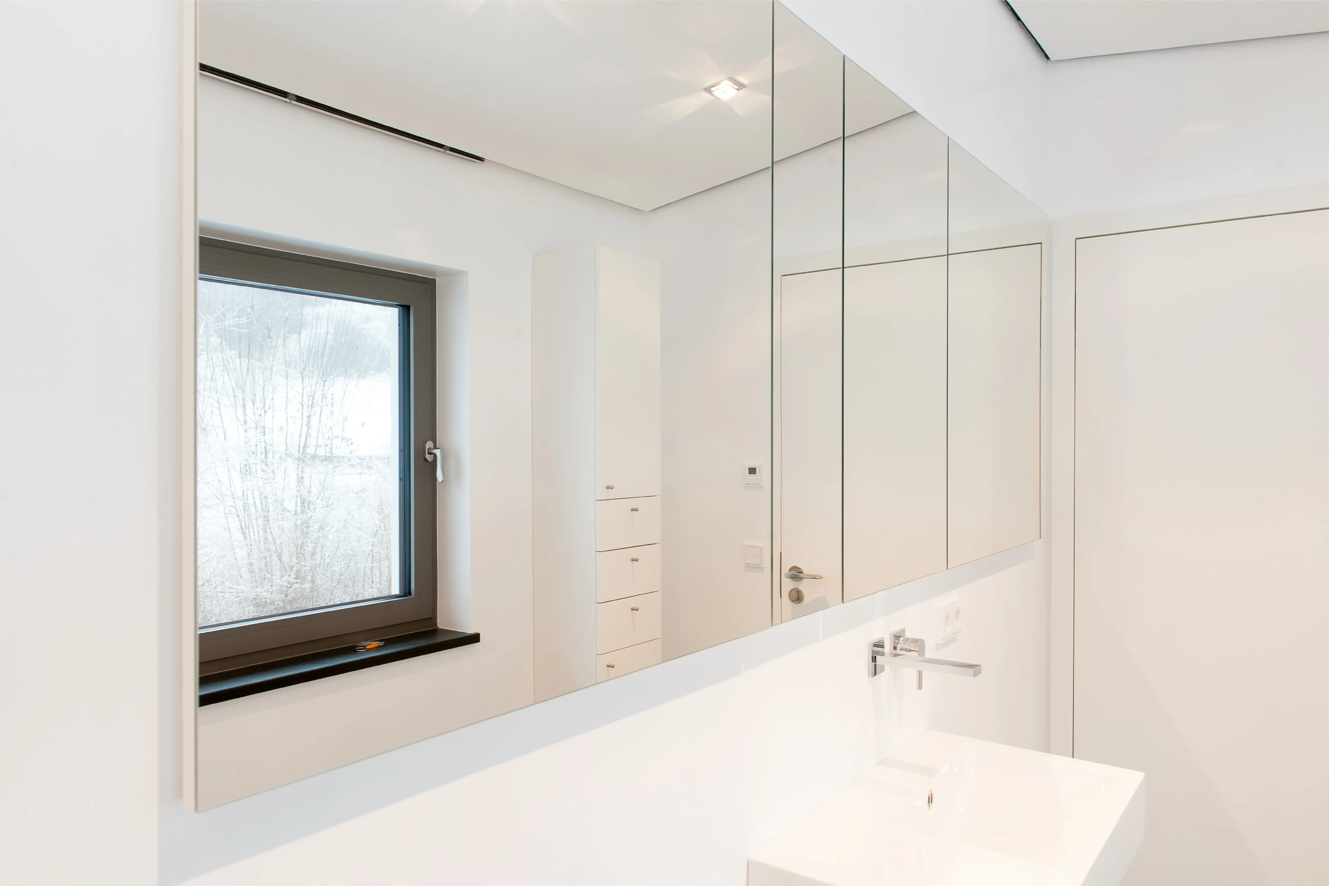 Modernes Badezimmer mit großer Spiegelfläche, weißem Waschtisch, Wandarmatur und Fenster ins Verschneite