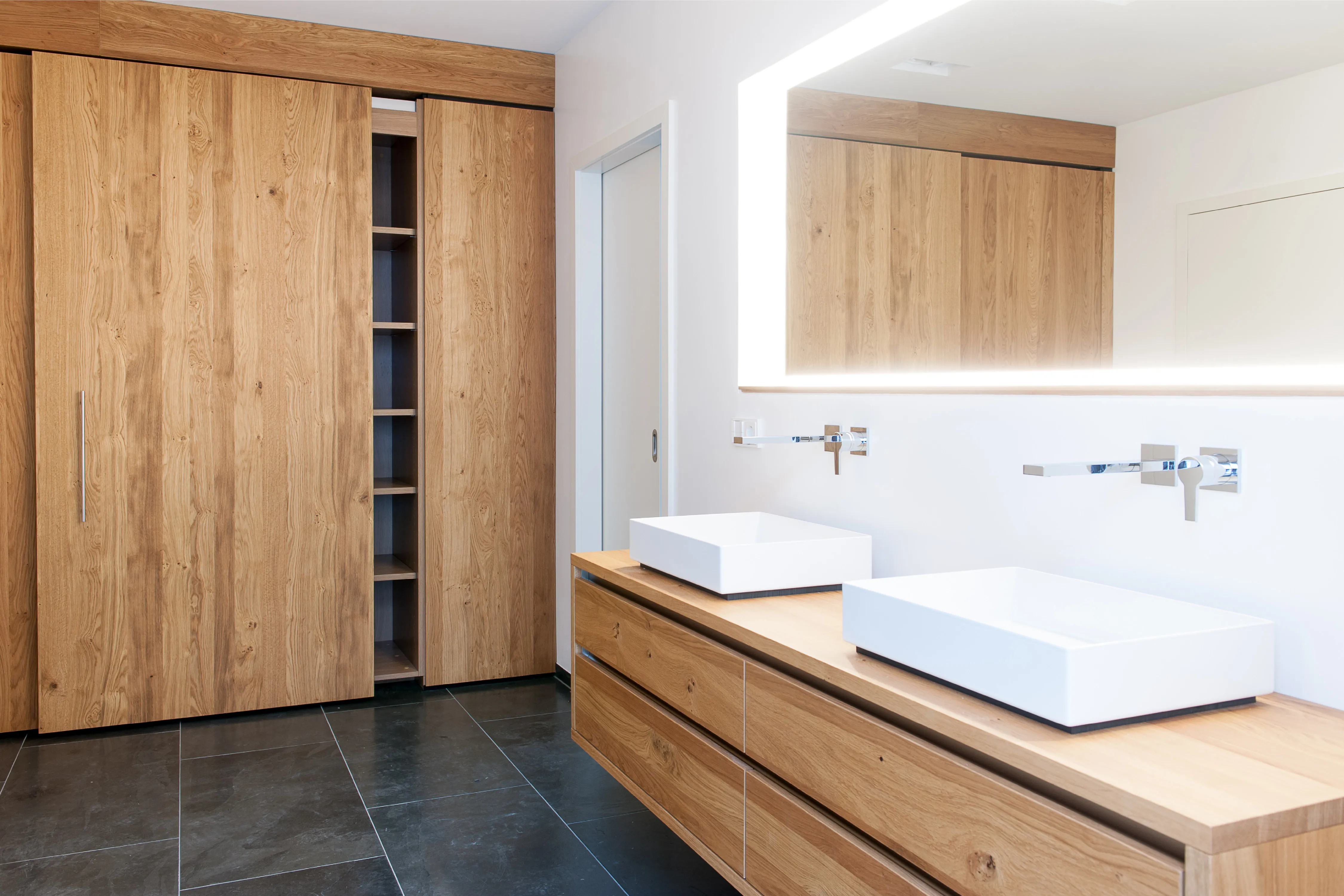 Badezimmer mit Doppelwaschtisch aus Holz, beleuchtetem Wandspiegel, dunklen Fliesen und Holzeinbauschränken