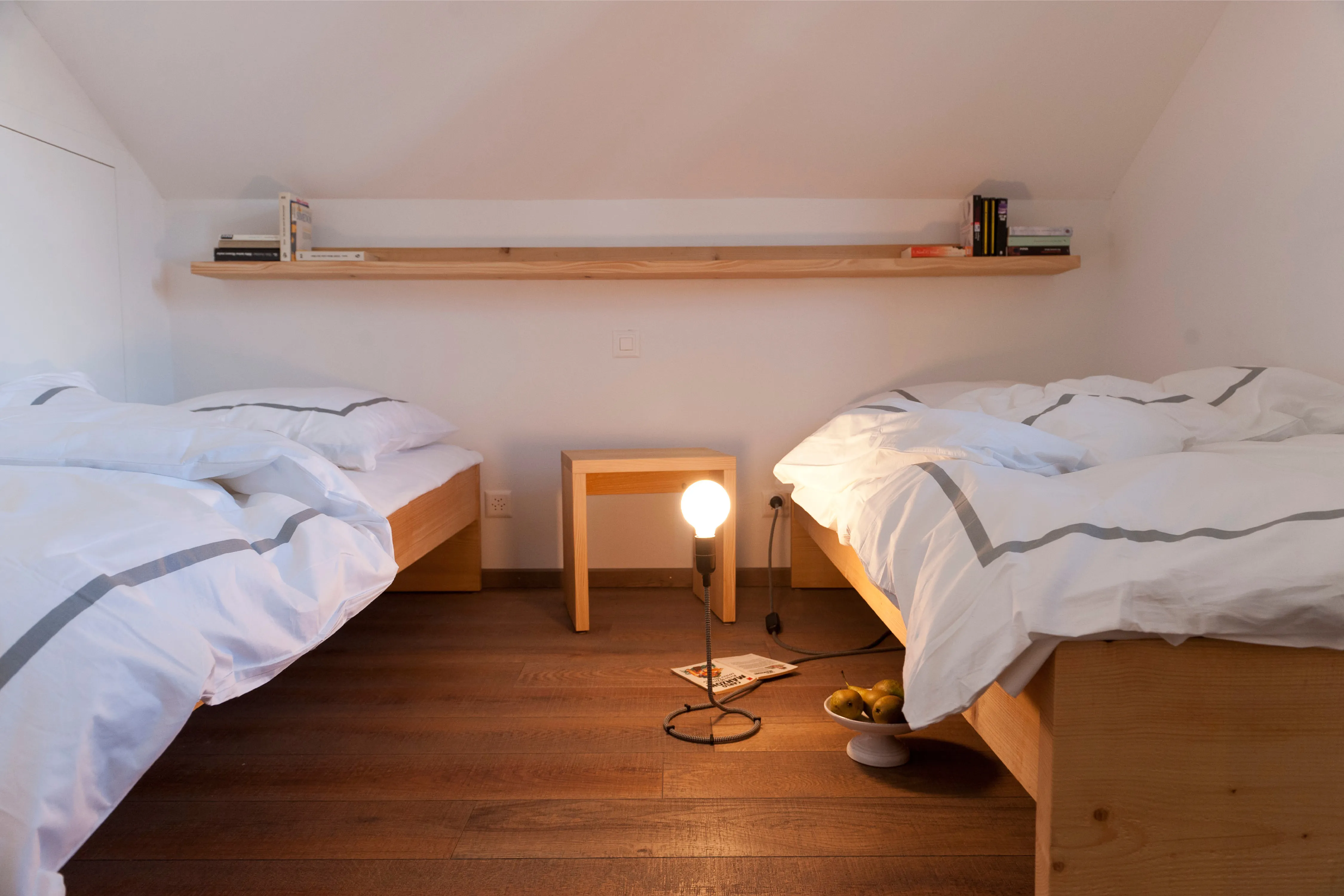 Gemütliches Schlafzimmer im Dachgeschoss mit zwei Einzelbetten aus Holz, Wandregal und einer Glühbirnen-Stehlampe am Boden