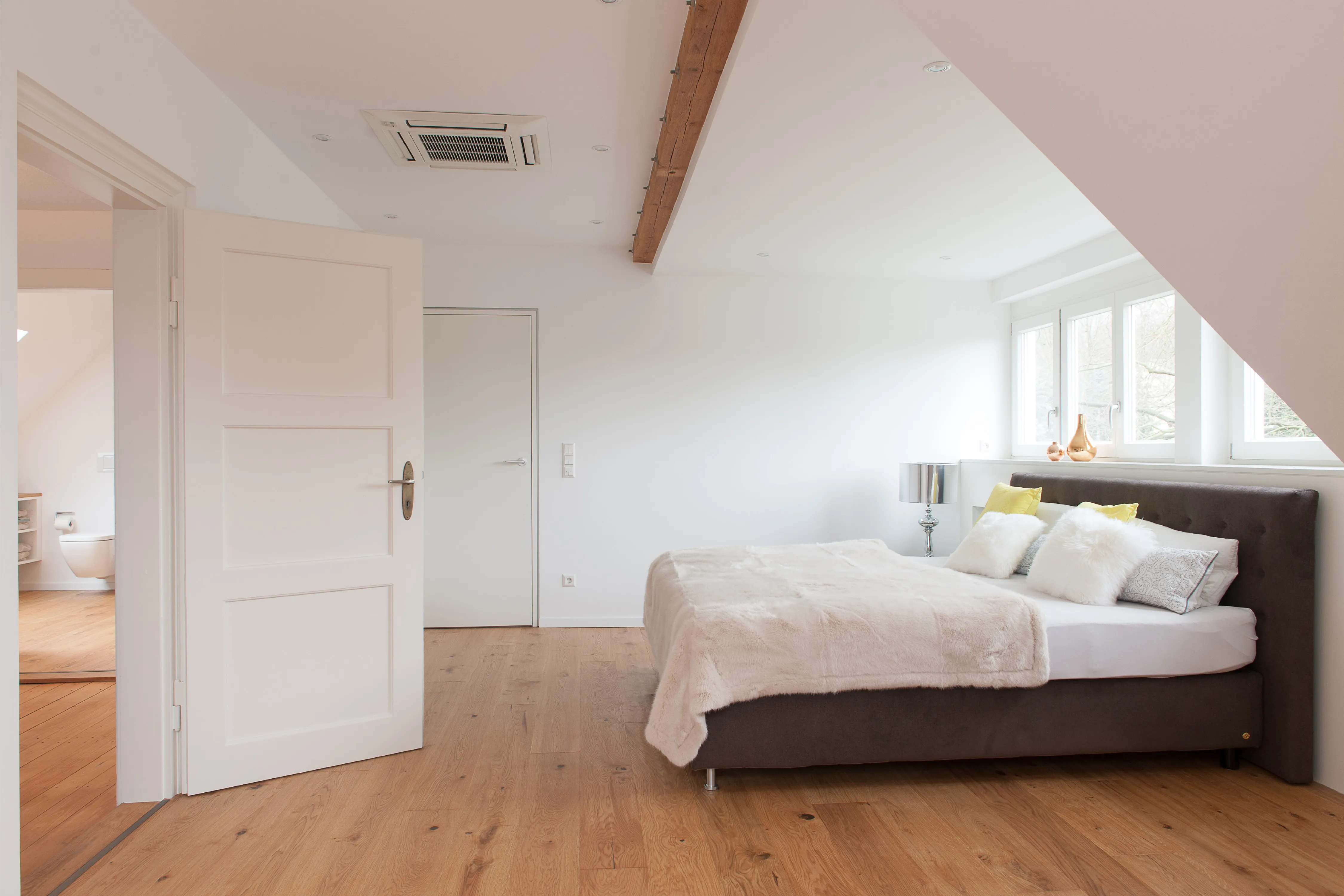 Helles Schlafzimmer unter dem Dach mit sichtbaren Holzbalken, offenem Holzboden, dunklem Boxspringbett mit hellem Fellüberwurf und Blick in das angrenzende Badezimmer.