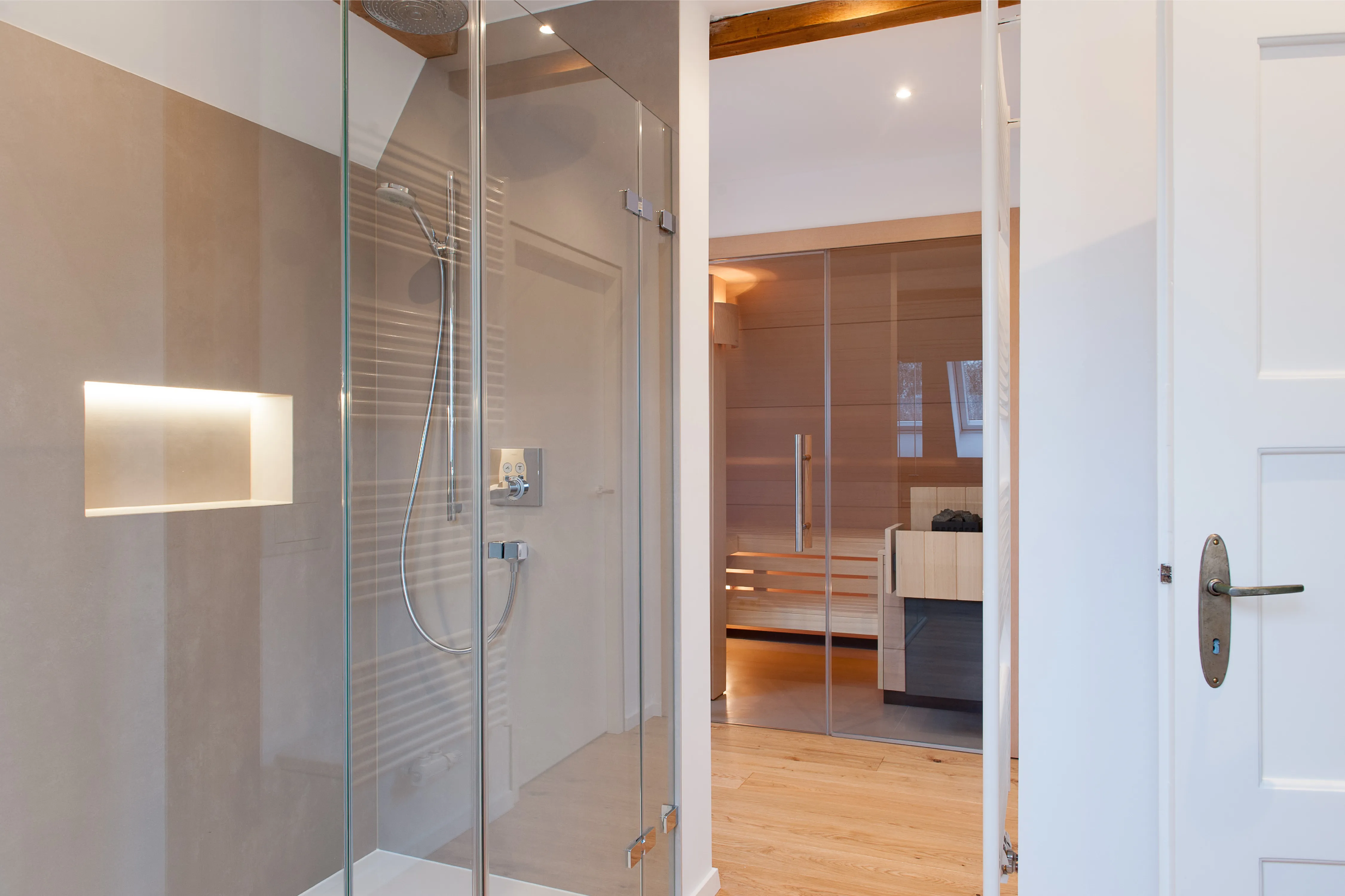 Blick im Badezimmer durch die verglaste Dusche mit beleuchteter Nische auf die angrenzende Wellness-Sauna.