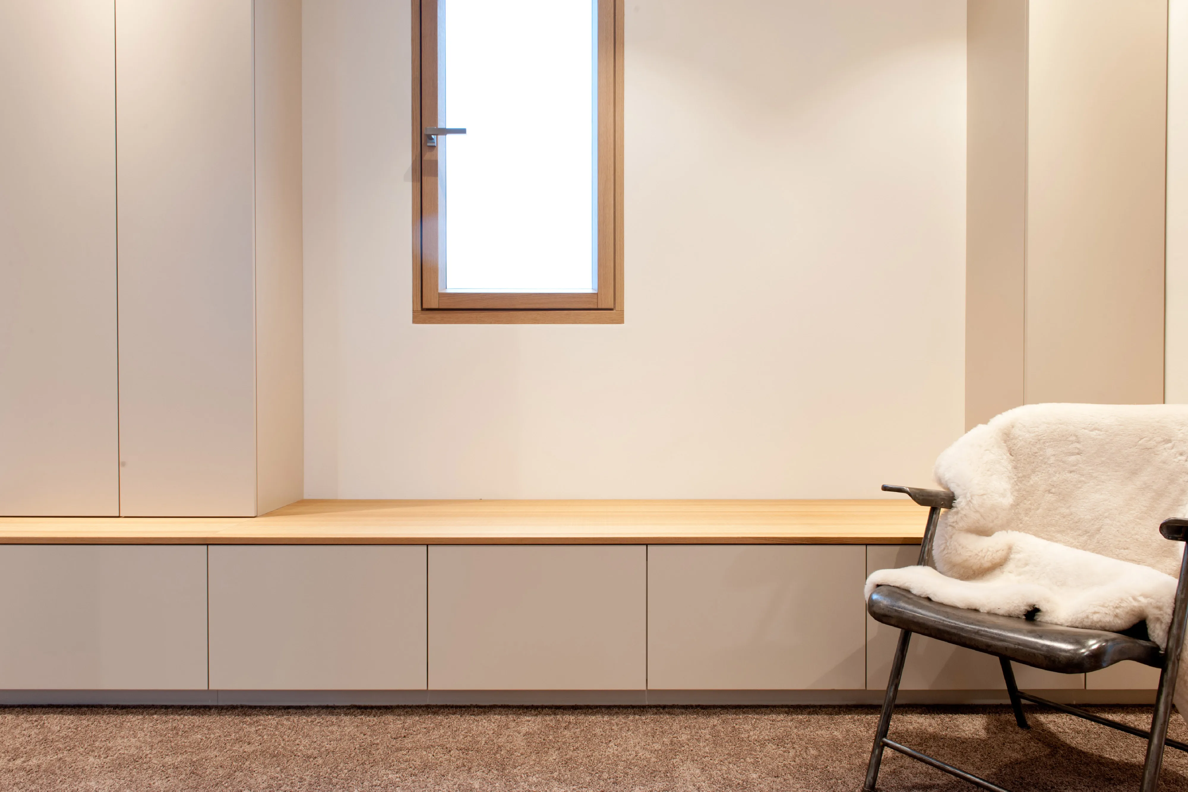 Maßgefertigte helle Sitzbank mit Holzabdeckung und Unterschränken neben einem Designstuhl mit Fellauflage