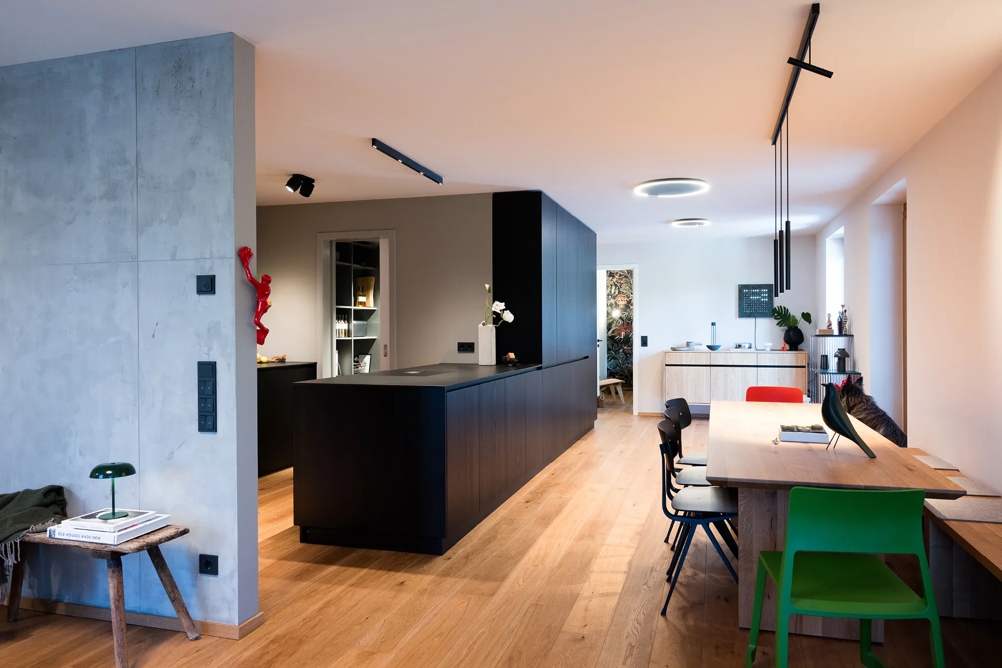 Offener Wohn- und Essbereich mit schwarzer Kochinsel, langem Holz-Esstisch und Wand in Betonoptik.