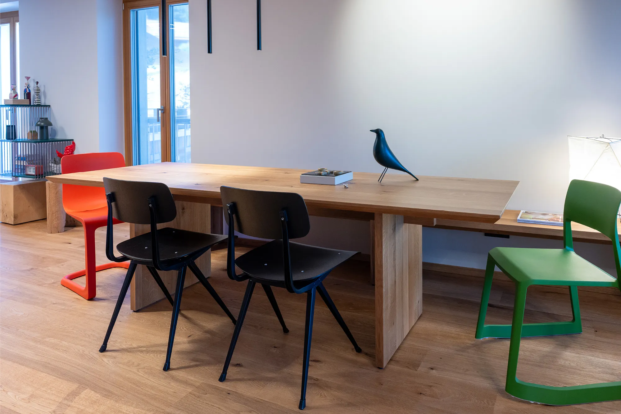Massiver Holz-Esstisch mit unterschiedlichen, bunten Designerstühlen und integrierter Sitzbank vor einer hellen Wand.