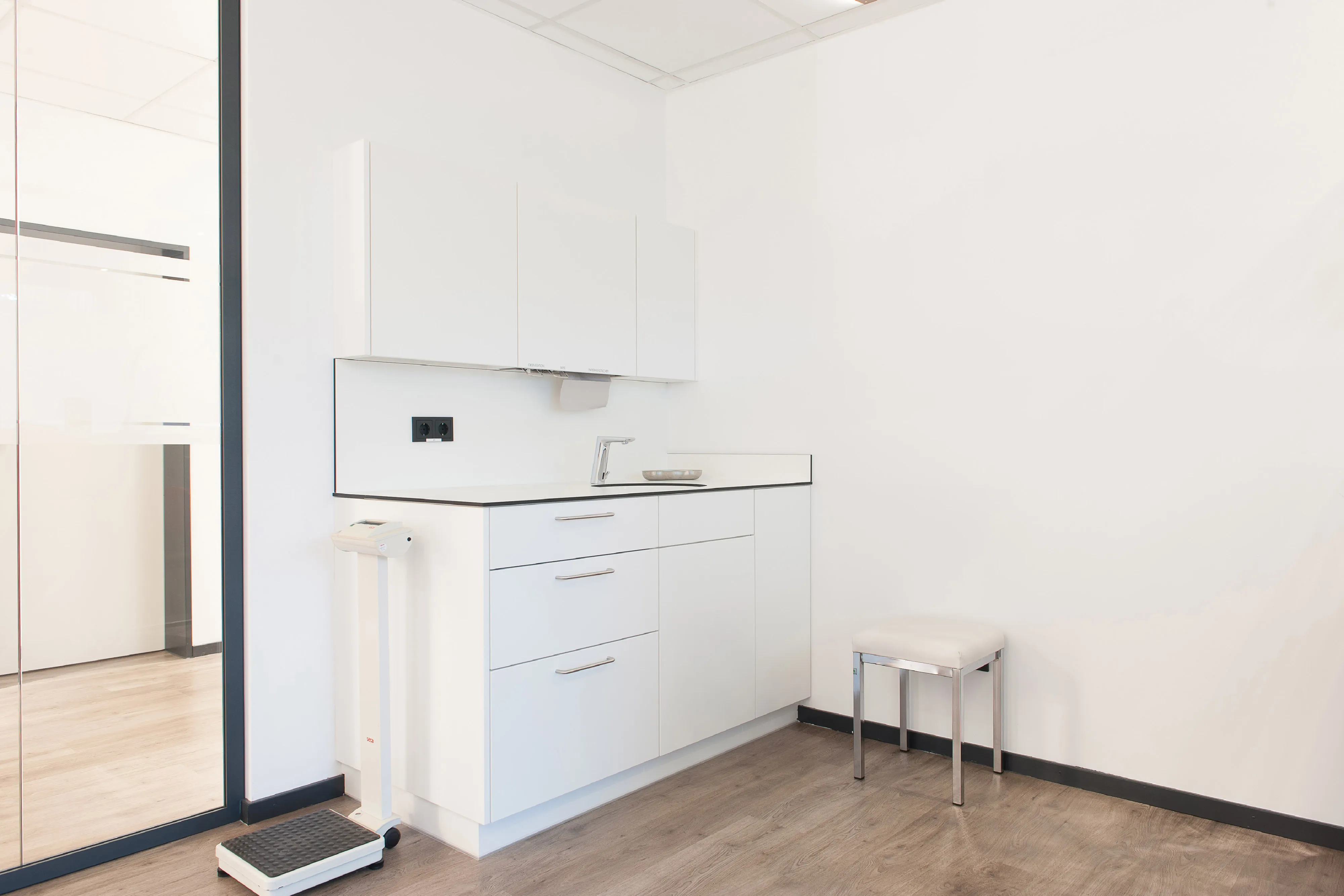 Behandlungsnische in der Praxis mit maßgefertigtem weißem Einbauschrank, Waschbecken und einer medizinischen Personenwaage.