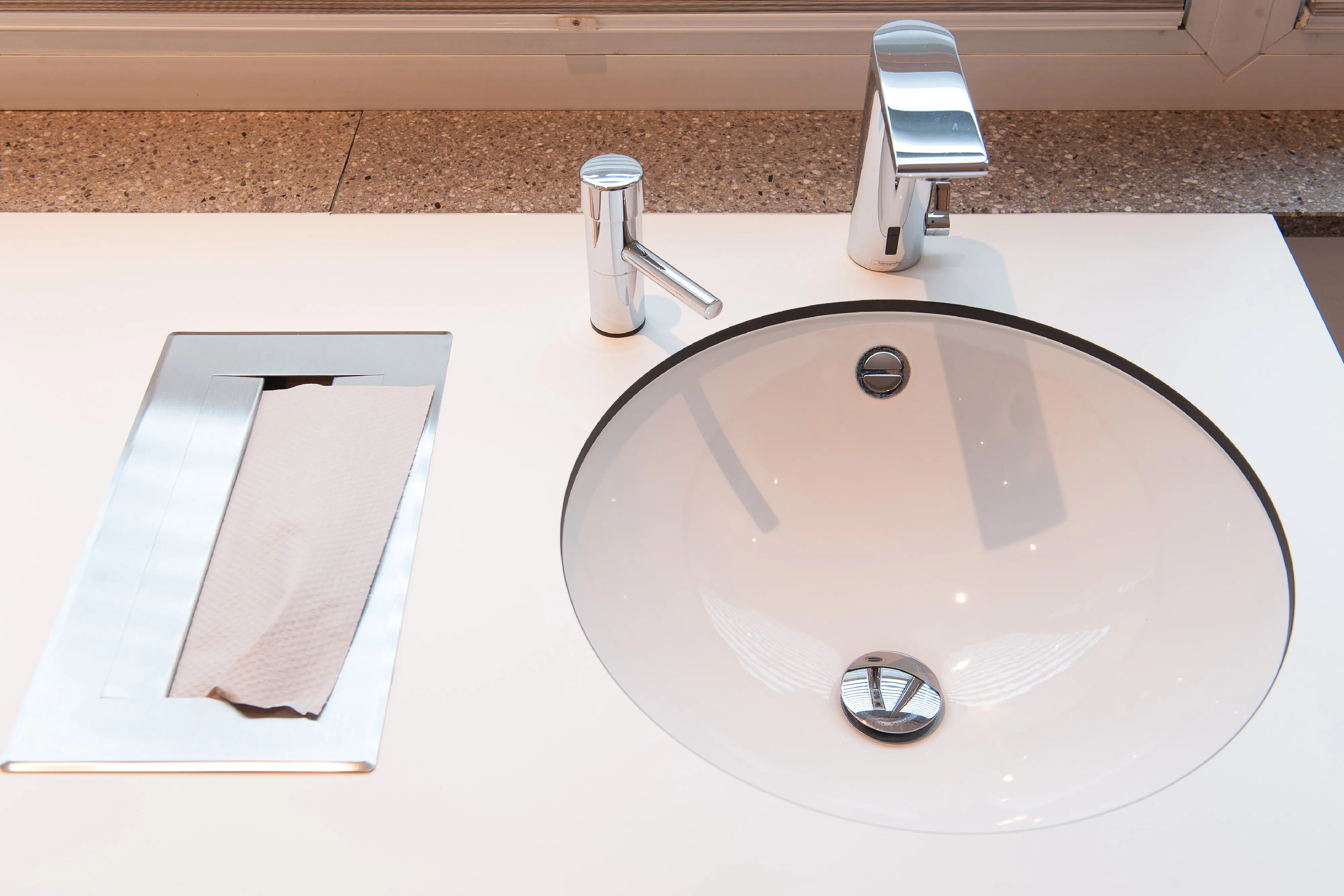 Detailansicht eines Waschtischs mit berührungslosem Wasserhahn, Seifenspender und flächenbündig verbautem Papiertuchspender.
