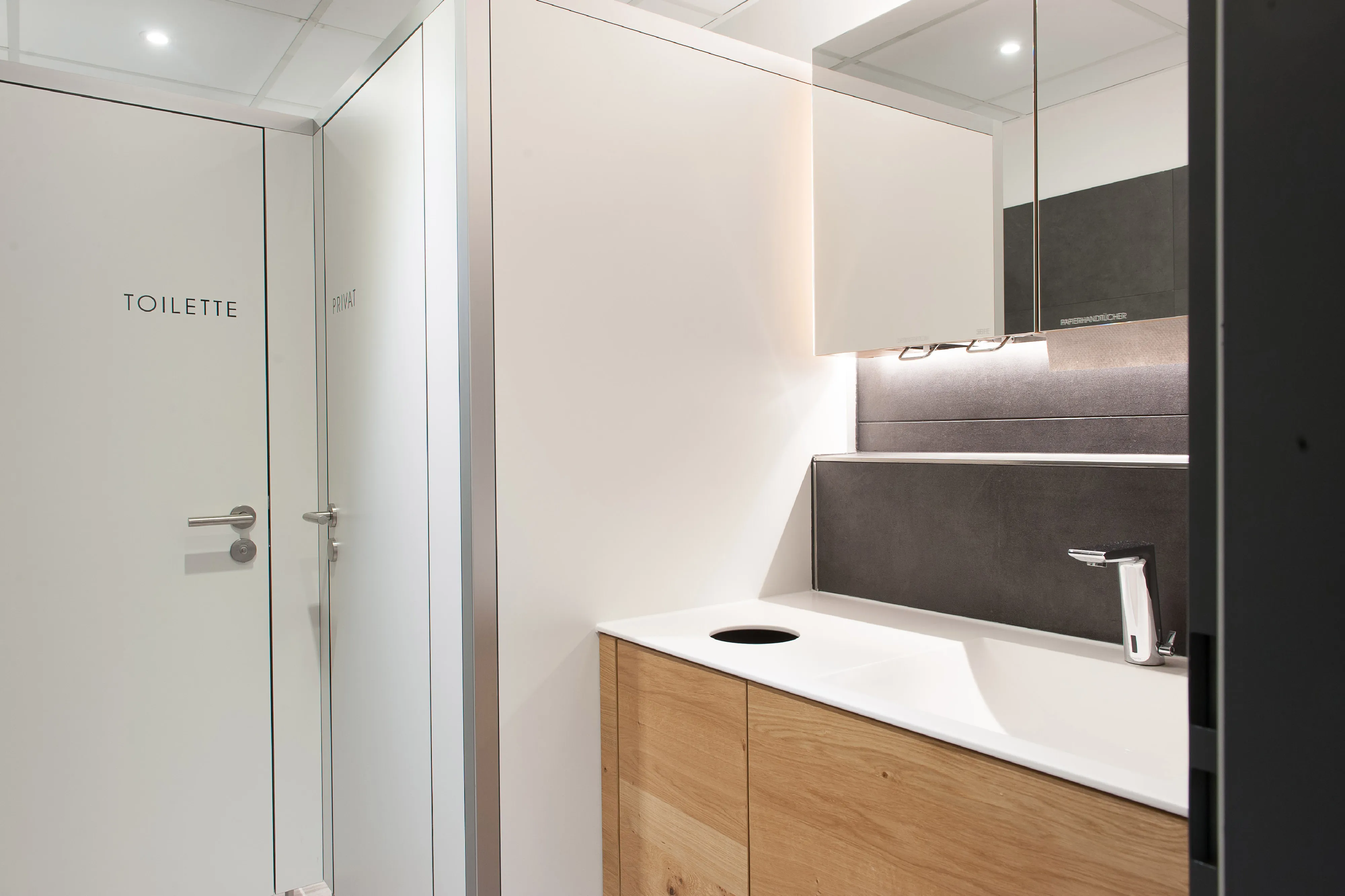 Patiententoilette mit Waschtisch aus Holz, beleuchtetem Spiegelschrank und blickdichten Kabinentüren.