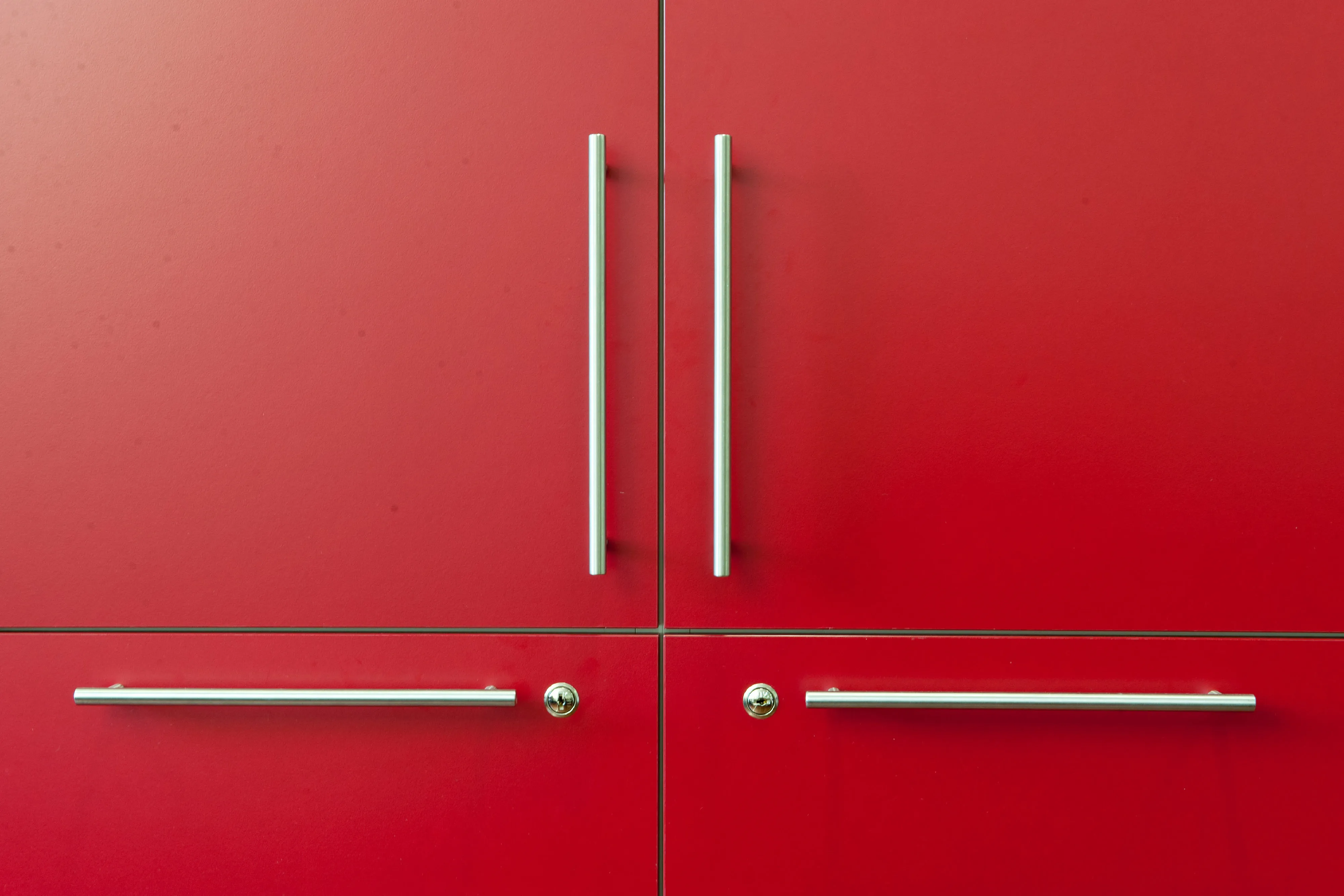 Nahaufnahme von vier Tür- bzw. Schubladengriffen in elegantem minimalistischen Design und passenden Schlosszylindern auf roter Schrankfront
