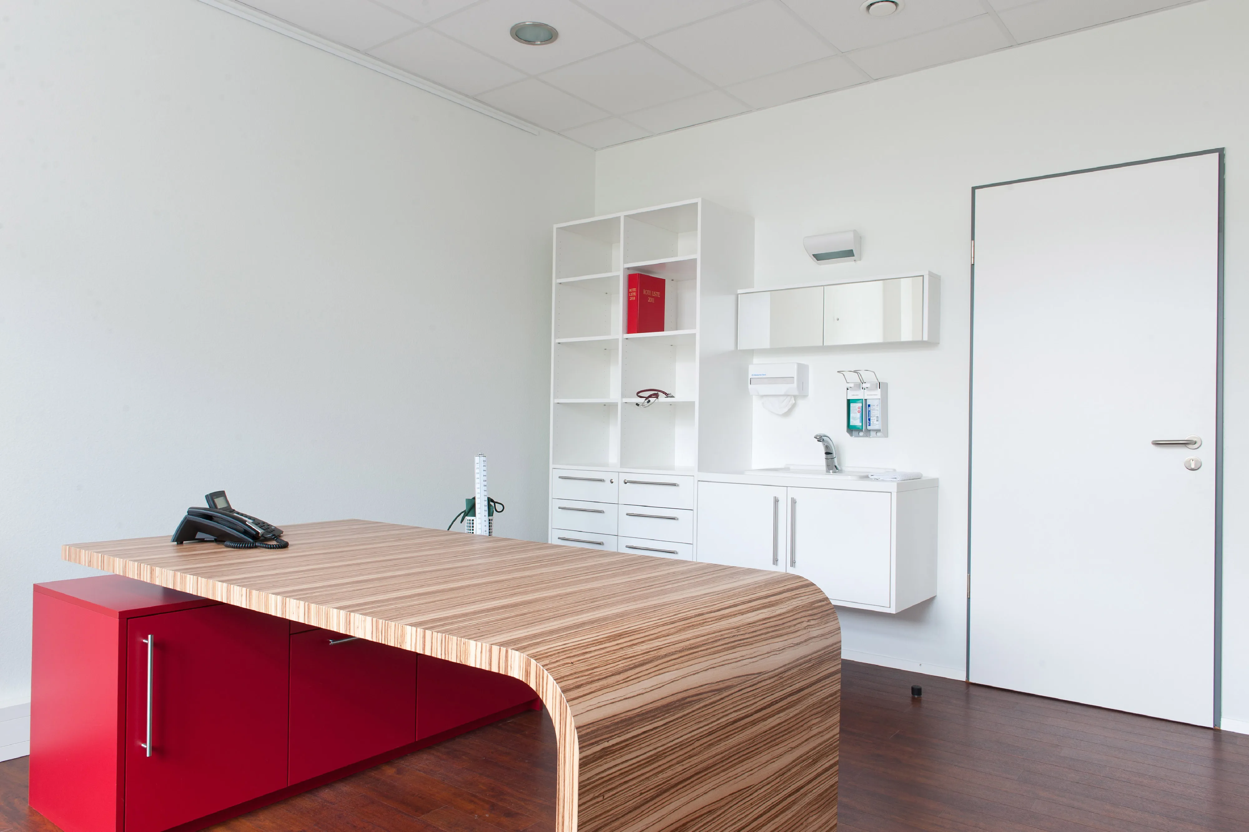 Behandlungsraum mit Tisch aus Vollholz in innovativem Design mit rotem Unterschrank, Regal und Waschbecken