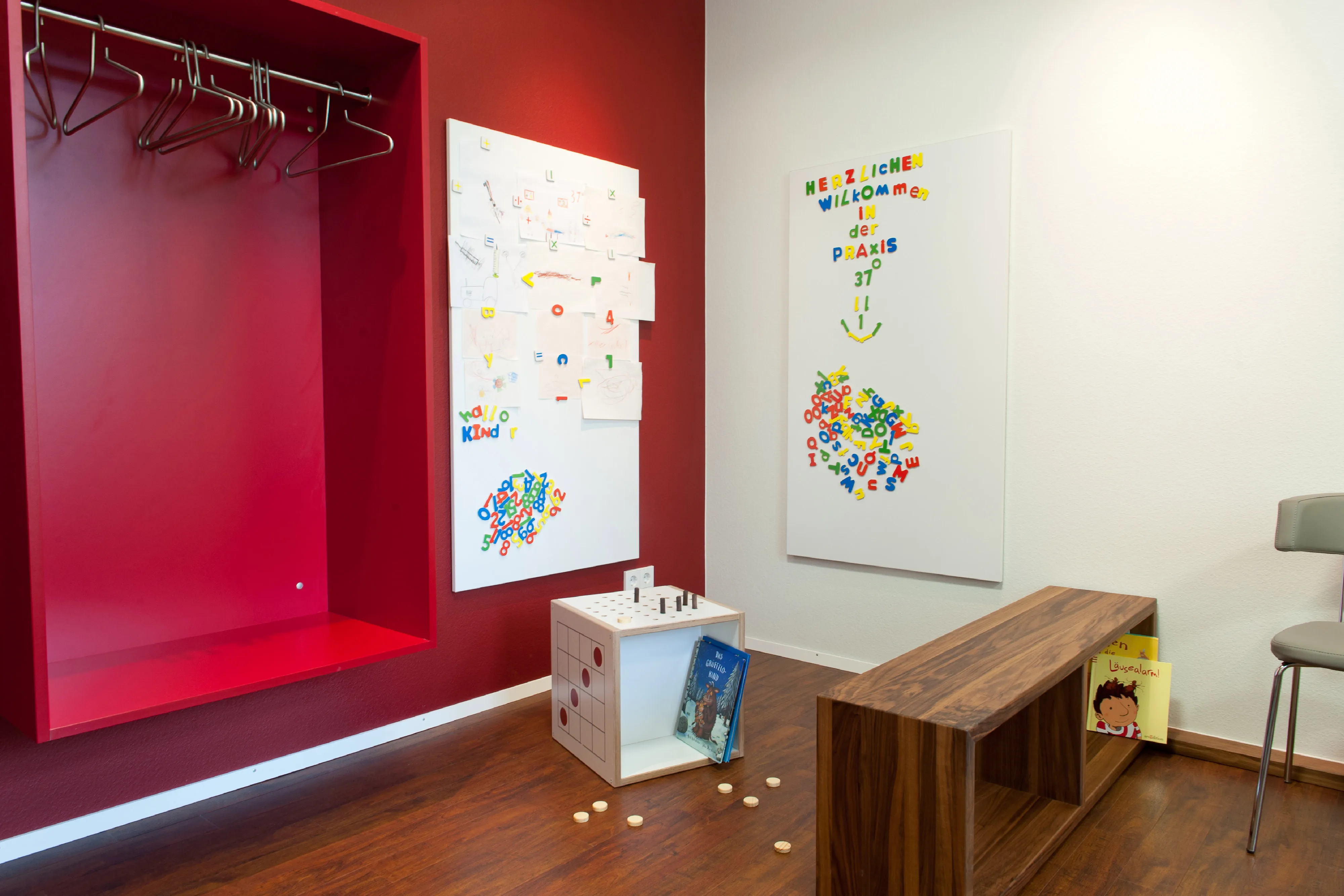 Wartezimmer mit Kinderecke. Farblich in die Wand integrierte rote Garderobennische, Magnettafeln mit Willkommensgruß und Holz-Sitzbank