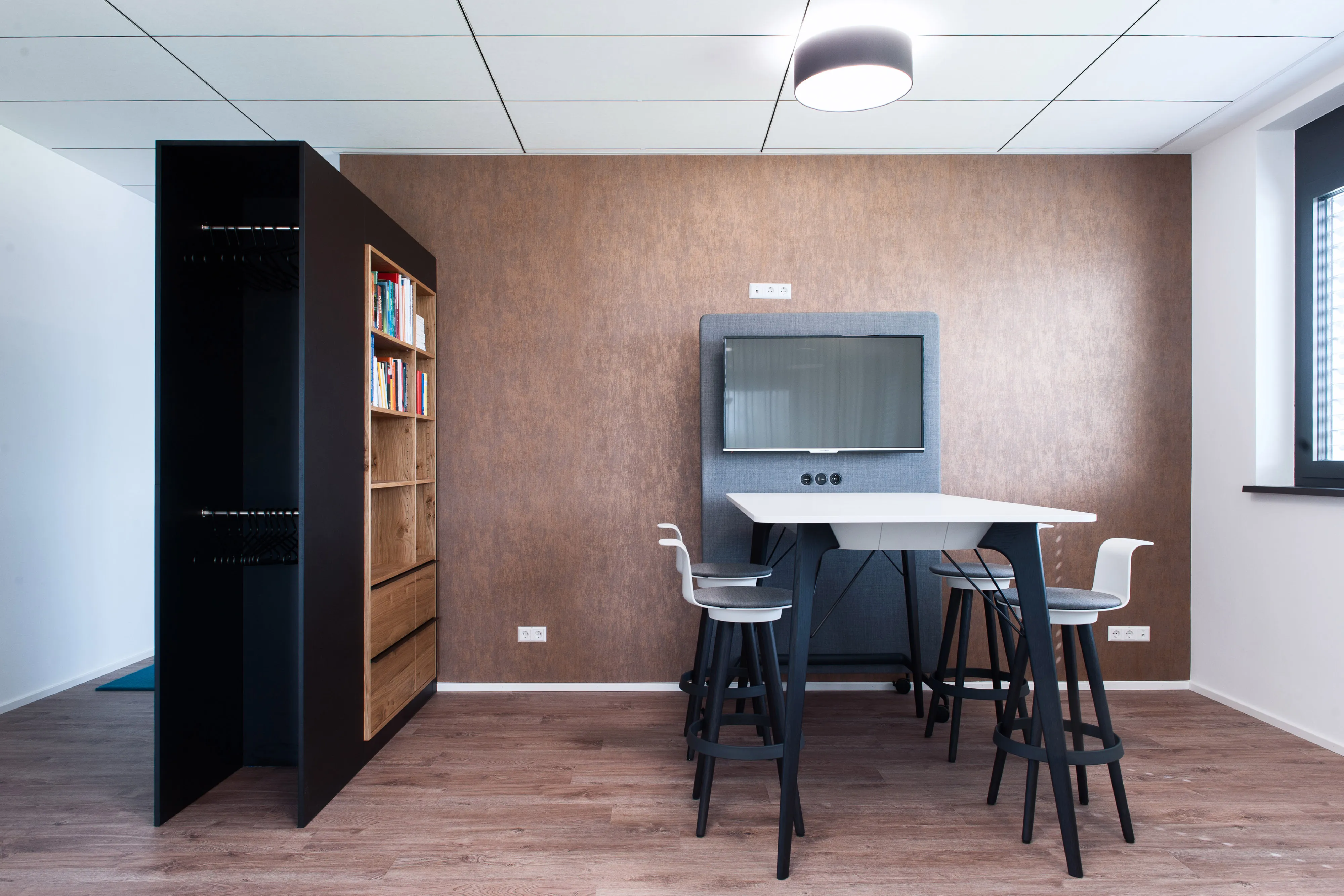 Besprechungsraum mit hohem Tisch, Barhockern, Bildschirm an der Wand und einem schwarzen Raumteiler mit integriertem Holzregal.