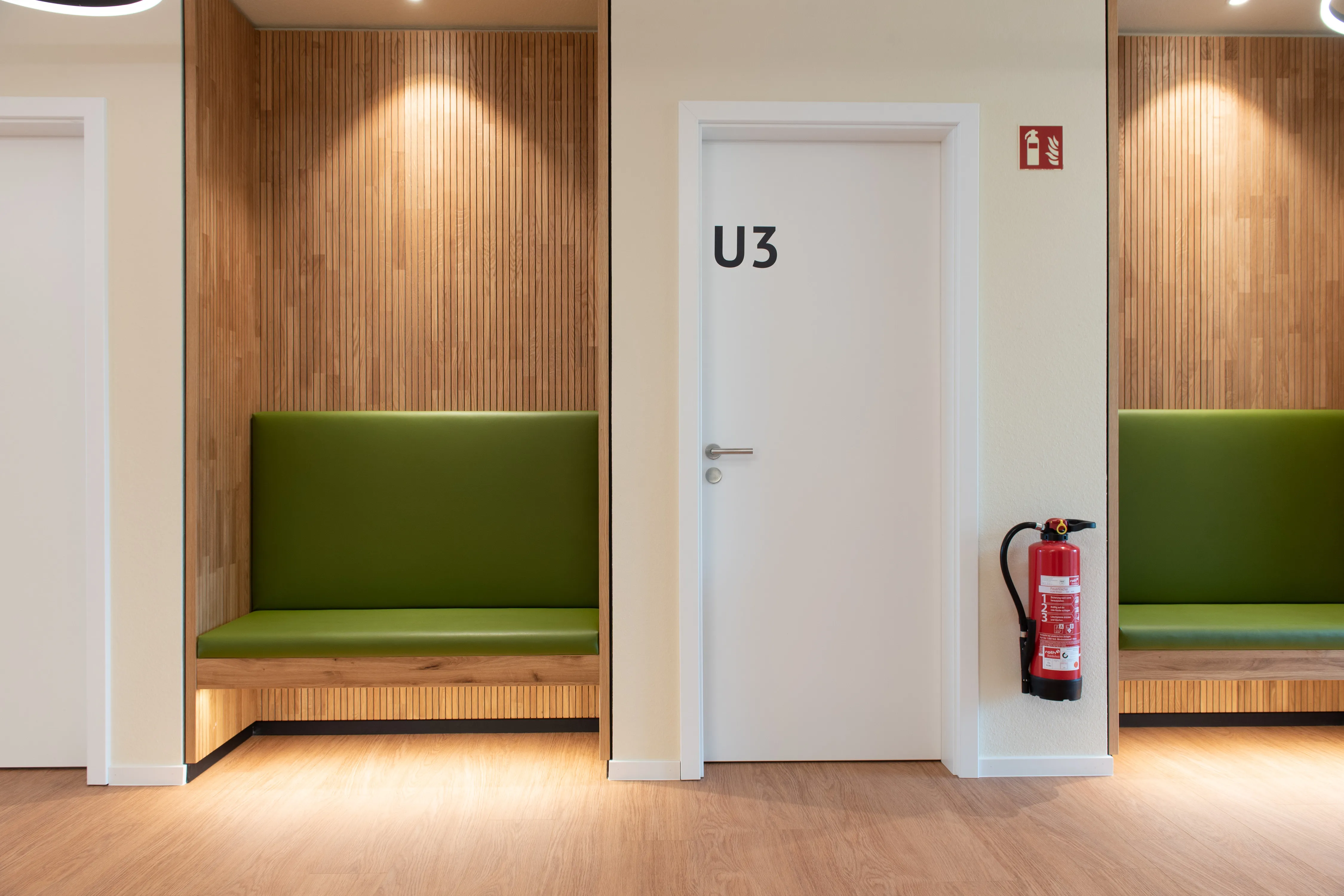 Frontalansicht einer passgenau eingebauten Warte-Nische mit holzverkleideter Rückwand und grün gepolsterter Sitzbank neben einer Praxistür.