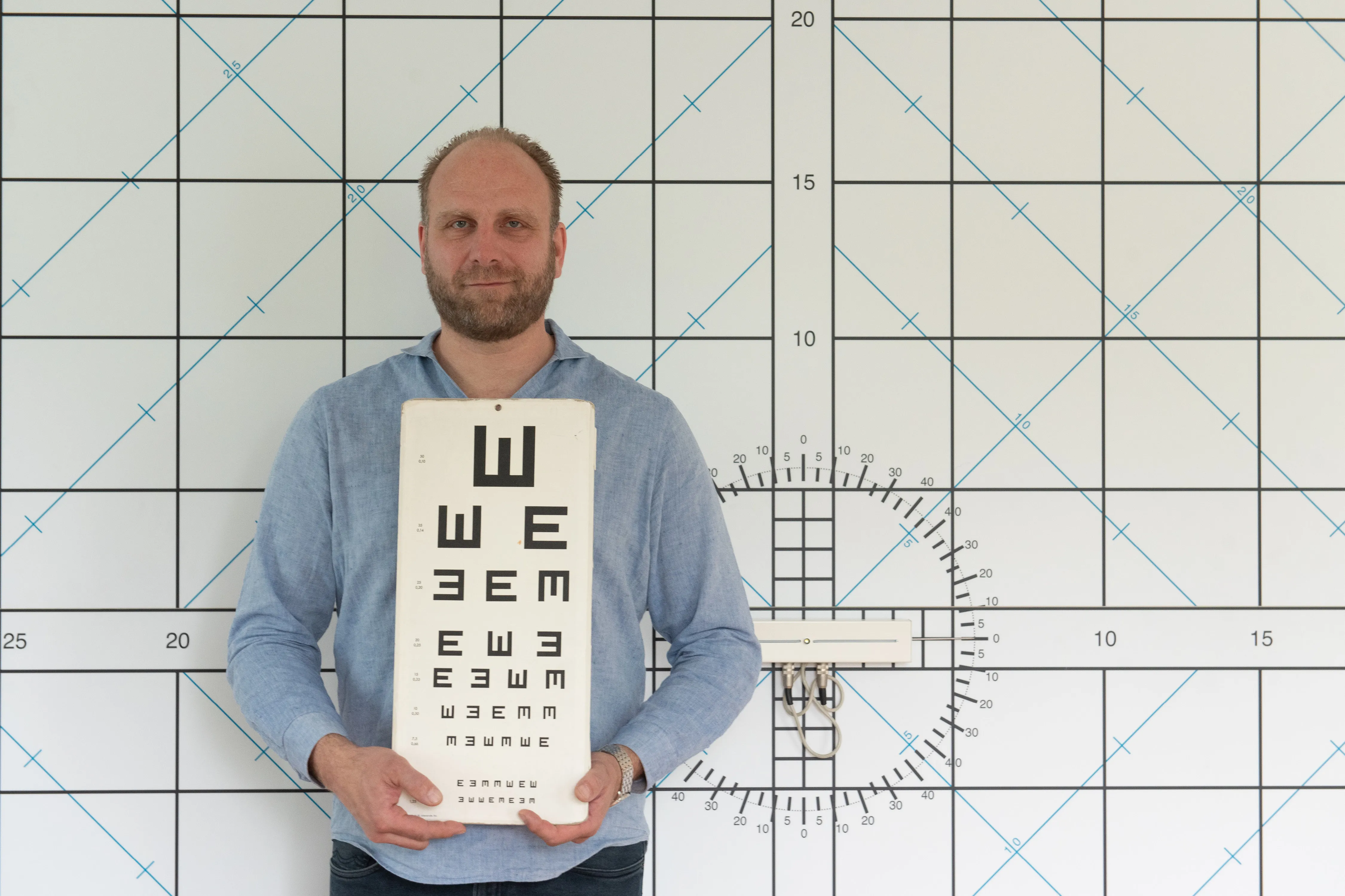 Bernhard Frankenhauser hält – im Verbrecherkartei-Style – die klassische Sehzeichentafel mit E-Haken vor einer großflächigen, an der Wand montierten Sehprüfungsskala.