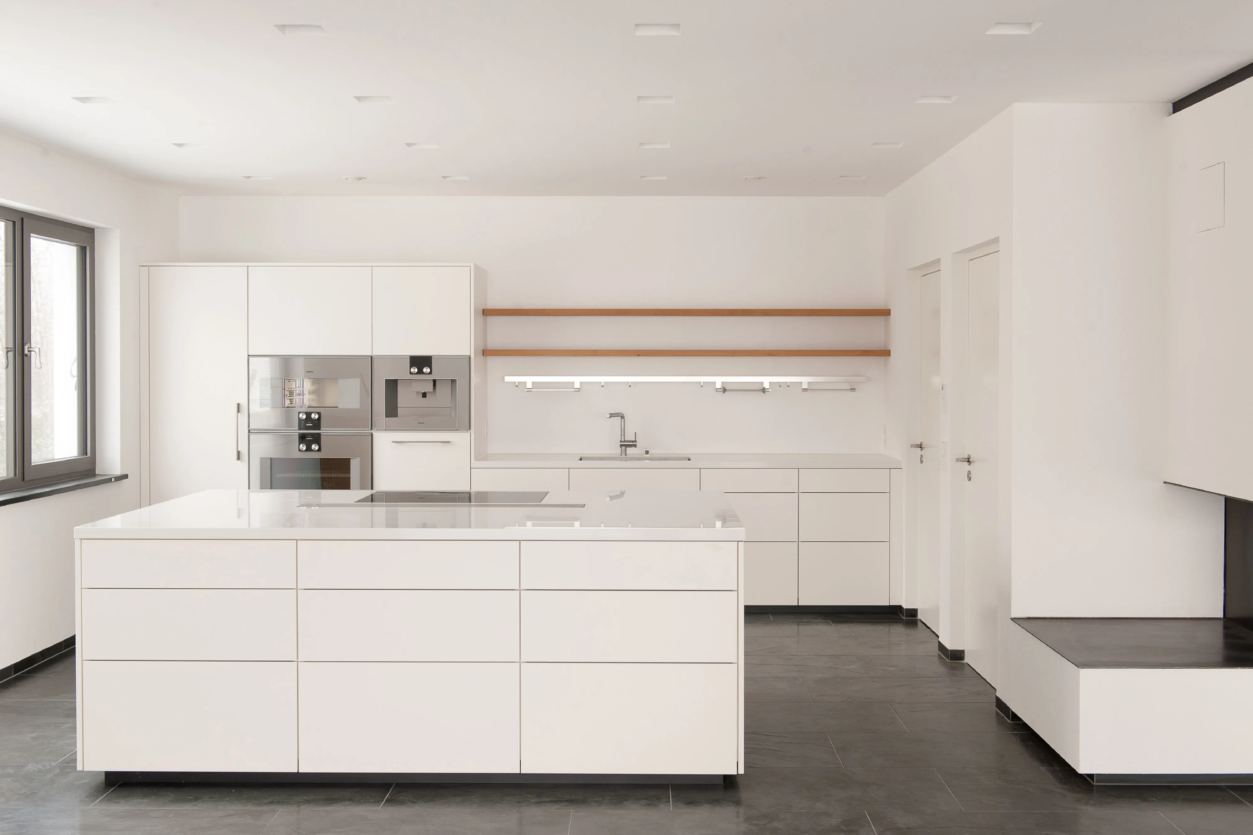 Großzügige, weiße Küche in minimalistischem Design mit Kochinsel, eingebauten Küchengeräten und dunklem Fliesenboden.