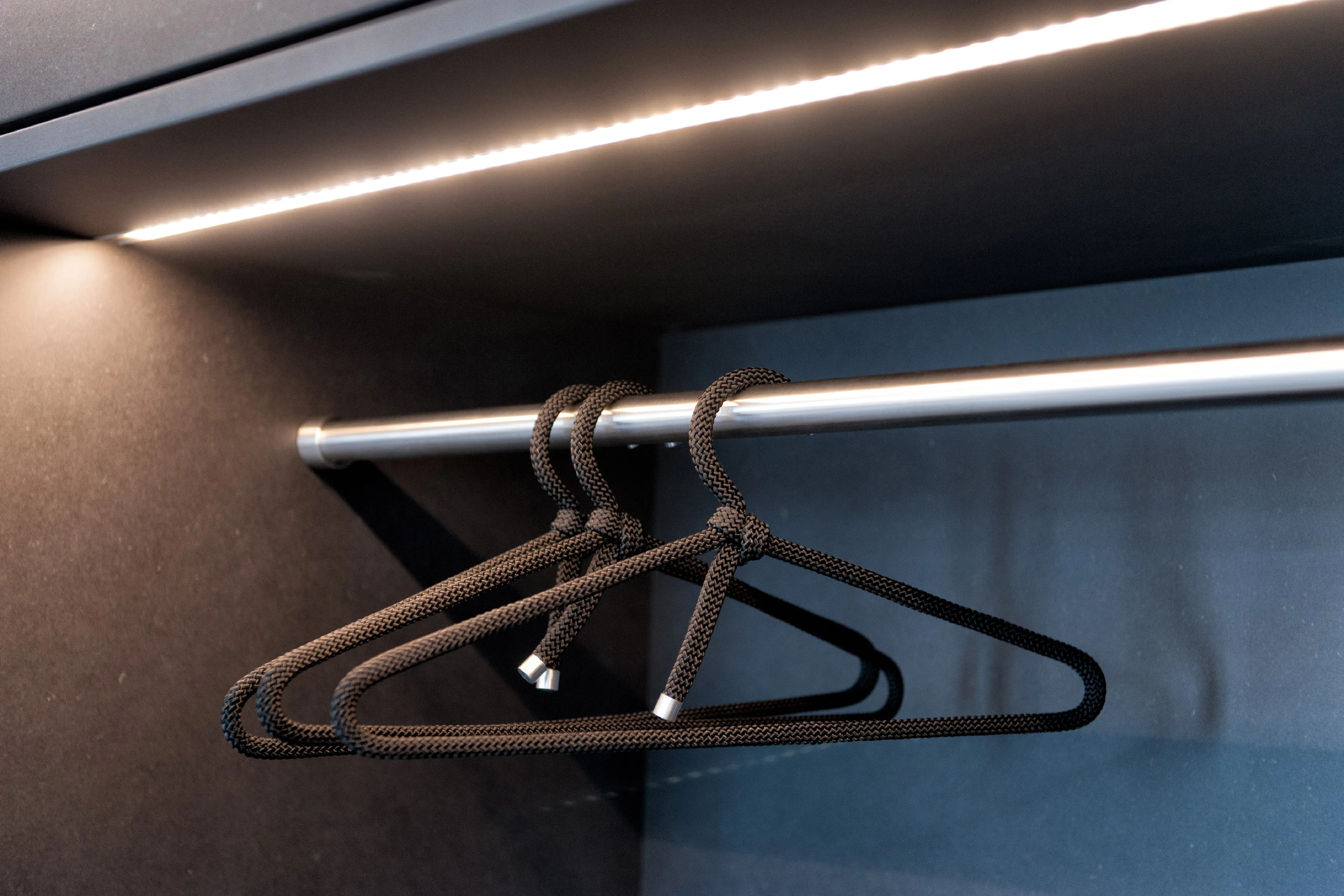 Detailaufnahme einer Edelstahl-Kleiderstange in einem dunklen Schrankkorpus mit schwarzer Kordel-Kleiderbügel-Garnitur und LED-Lichtleiste.