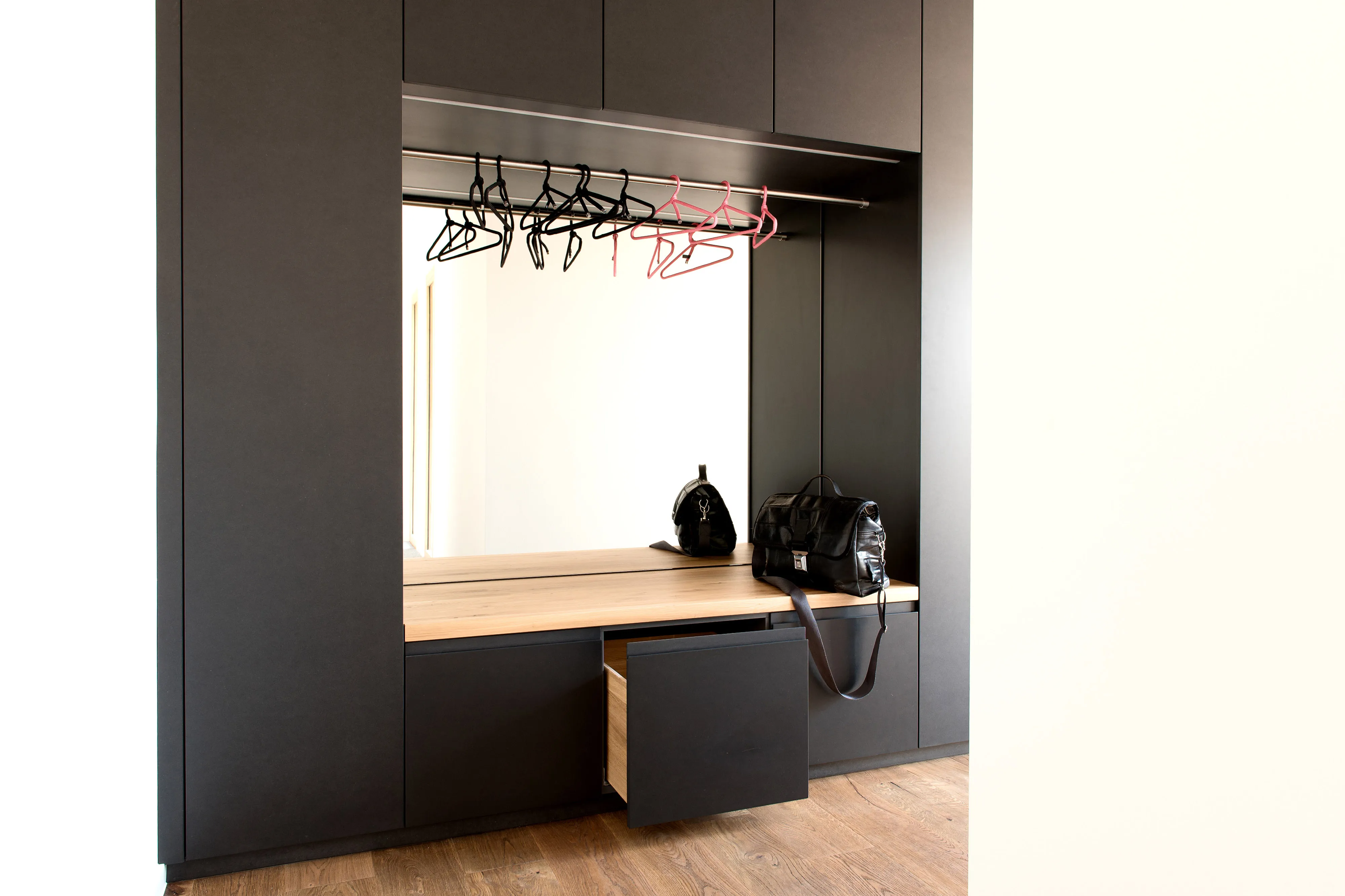 Schwarze Einbau-Garderobe mit Eichenholz-Sitzfläche, großer Spiegel-Rückwand und einer geöffneten, bodennahen Schublade.