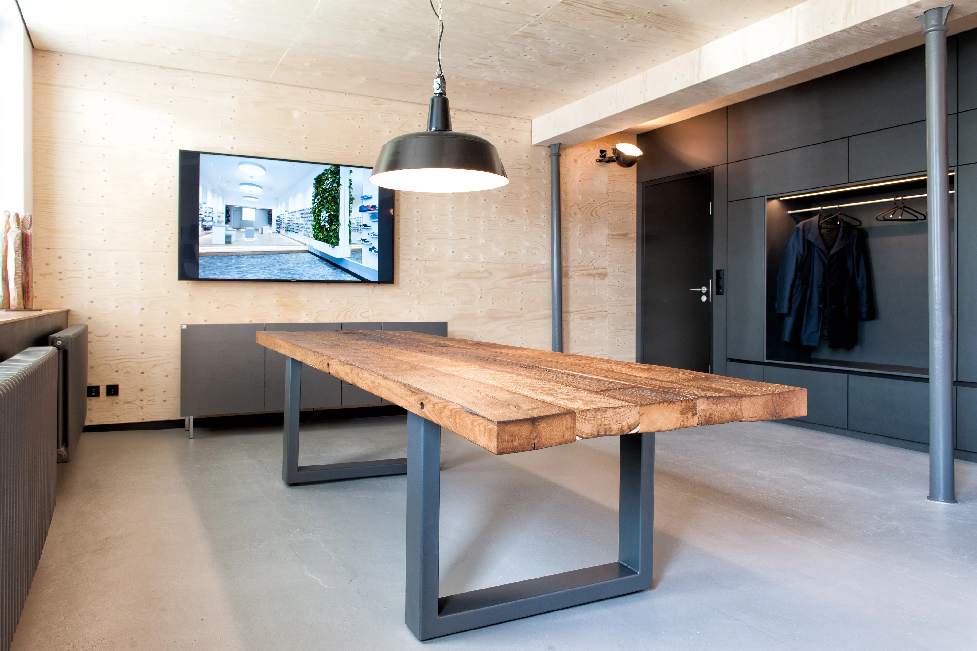 Rustikaler Massivholztisch aus urigen Holzbohlen auf grauen Metallkufen in einem modernen Ausstellungsraum mit Holz-Wandverkleidung.