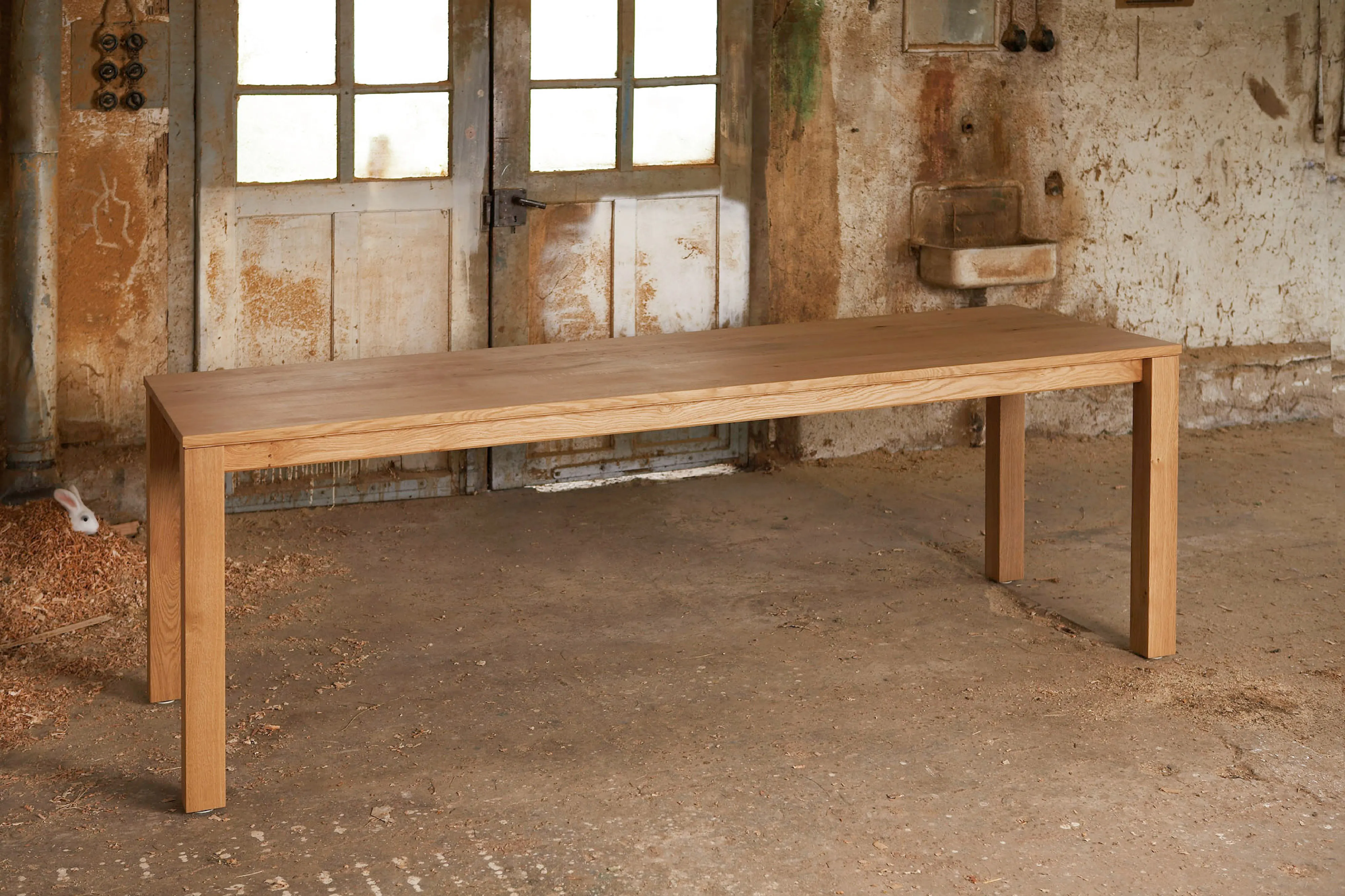 Schlanker, klassischer Esstisch aus hellem Massivholz mit vier Ecken-Tischbeinen vor historischem Werkstatt-Hintergrund.