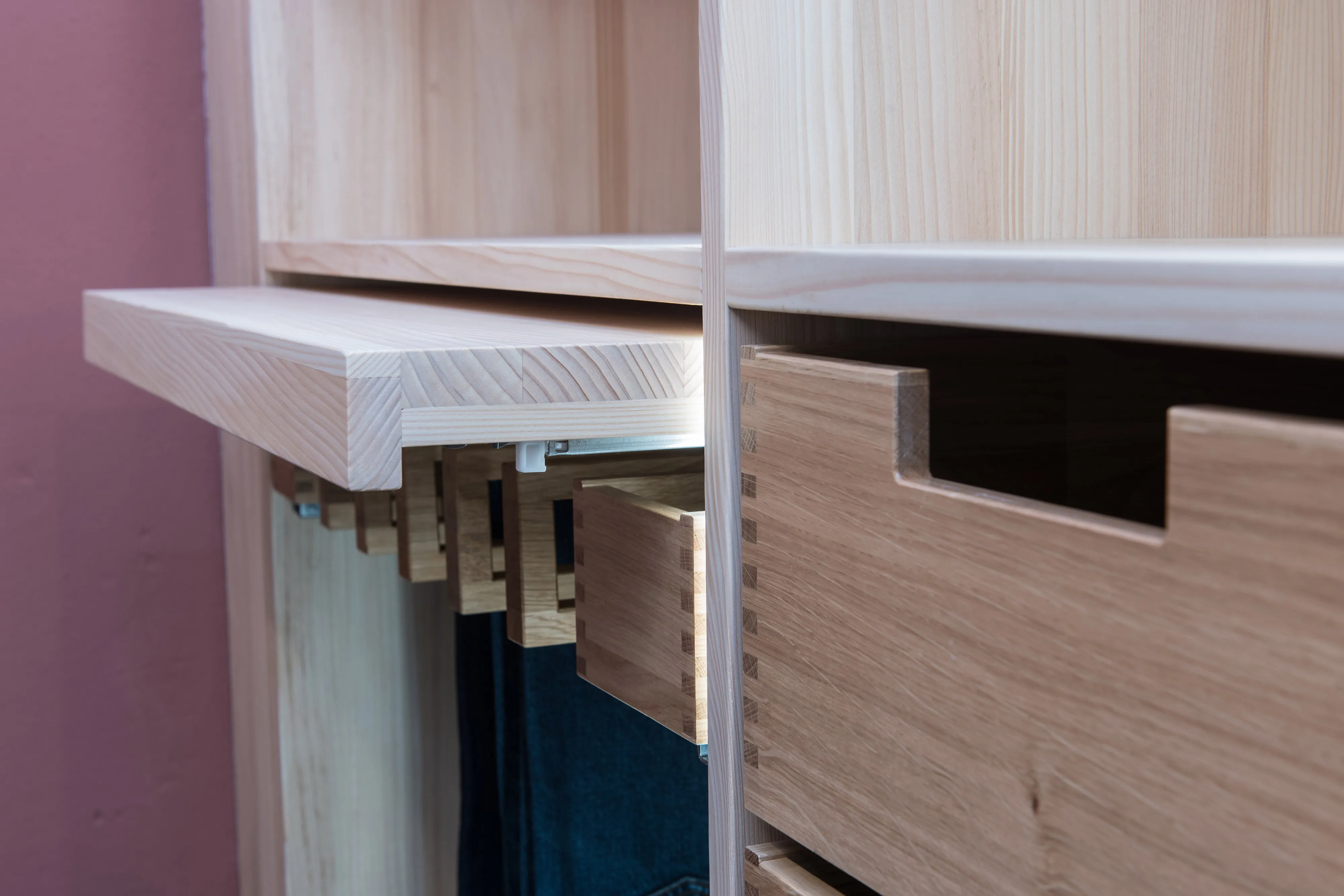 
Detailansicht eines offenen Kleiderschranks aus hellem Holz mit beleuchtetem Auszug und handgefertigter Holzschublade.