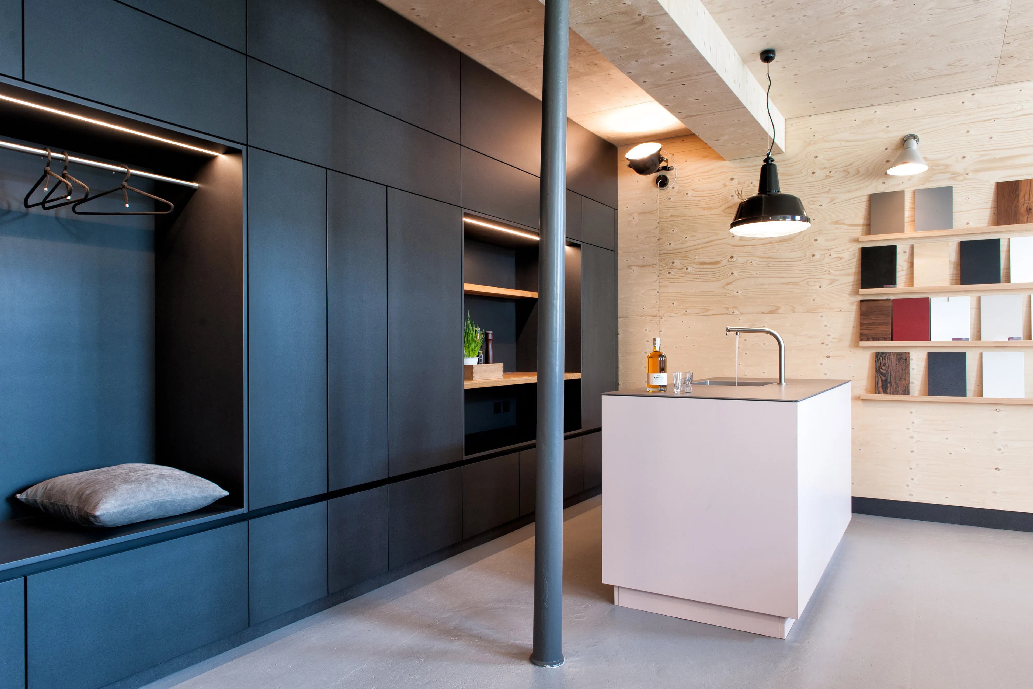 Besprechungszimmer mit Materialmustern, schwarzer Einbau-Garderobe, und hellem Küchen-Arbeitsblock.