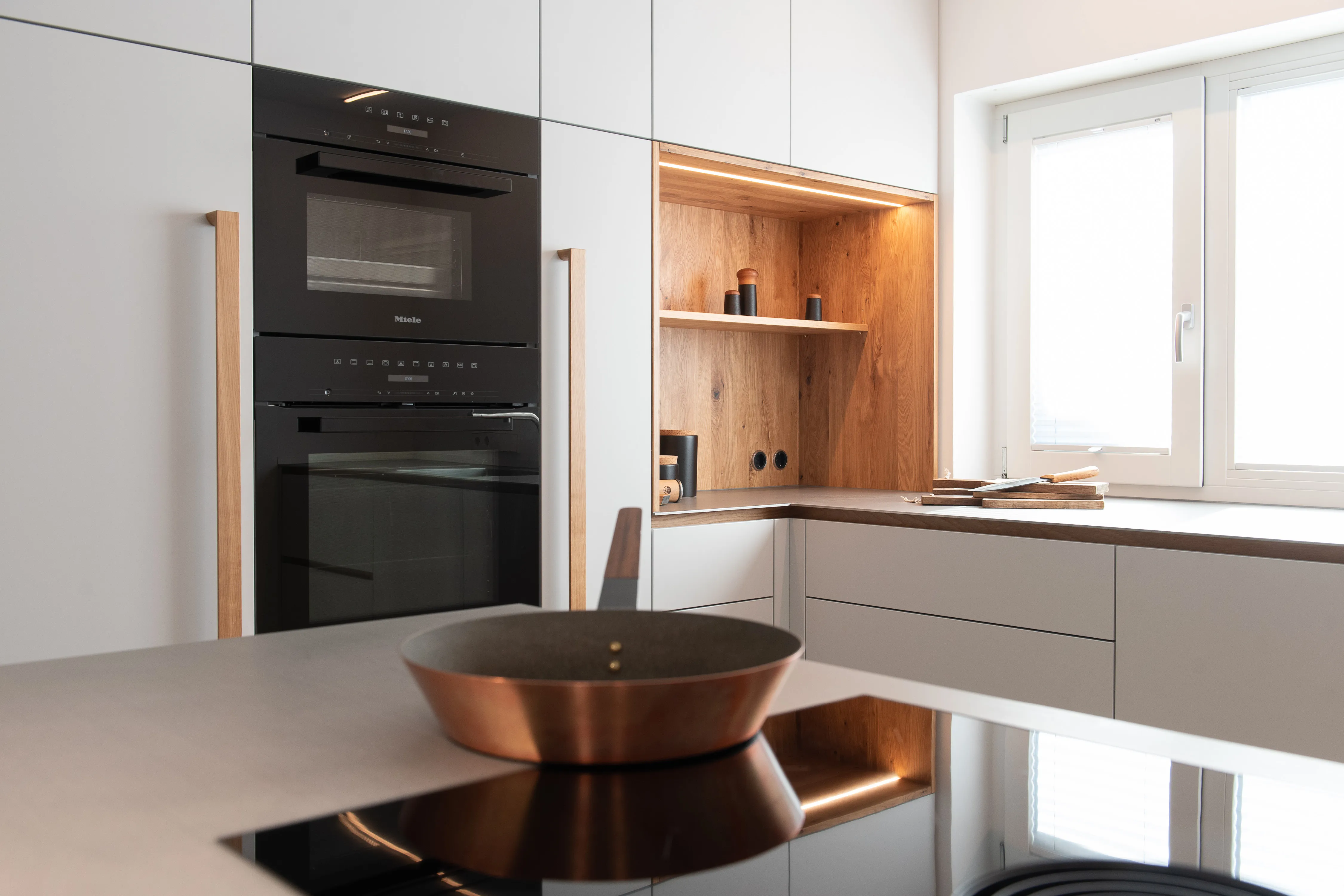 Detailansicht einer hellen Einbauküche mit Miele-Backofen, Kochgeschirr im Vordergrund und beleuchteter Echtholz-Nische.