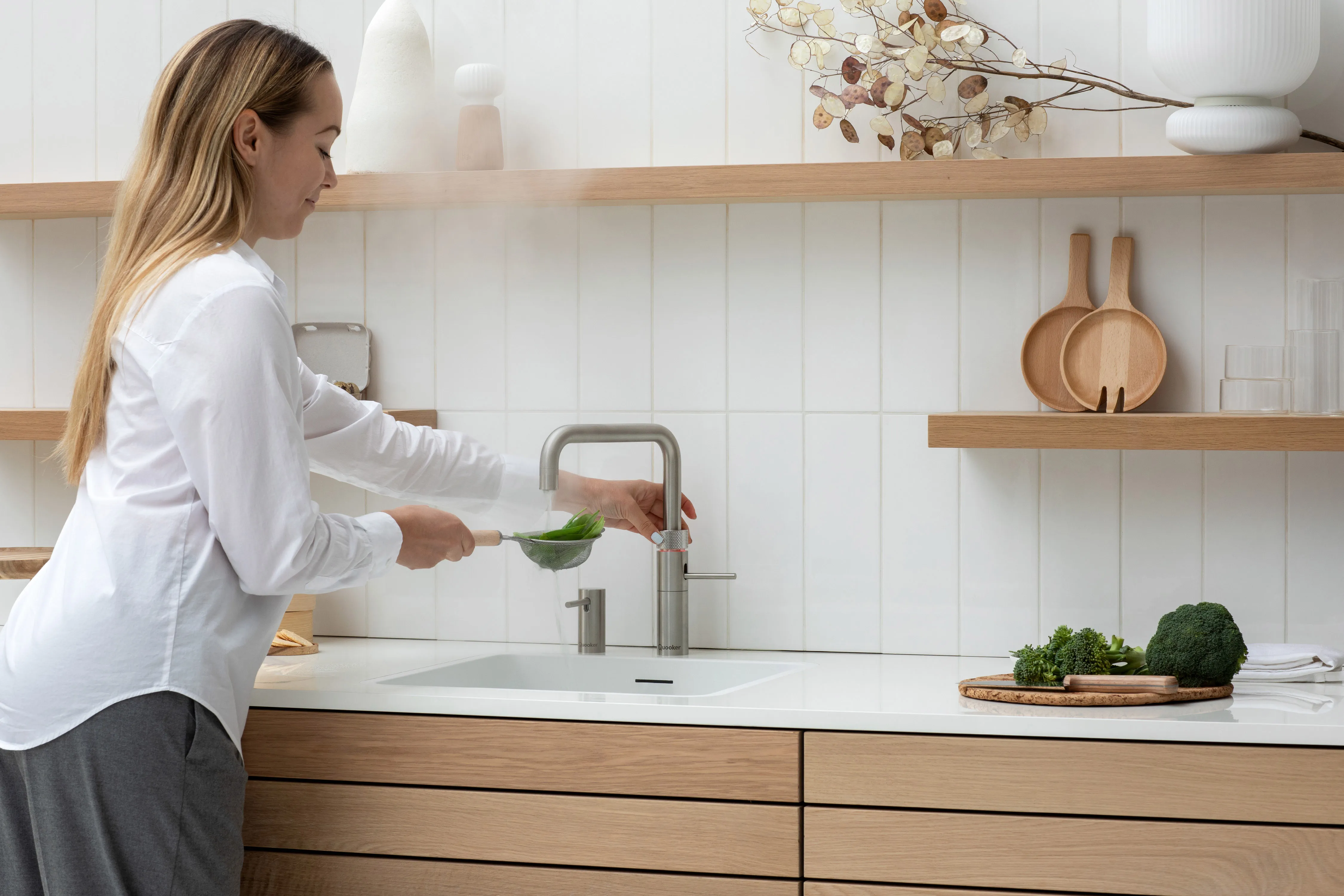 Frau blanchiert Gemüse mit kochendem Wasser aus einem Quooker-Edelstahl-Wasserhahn in einer hellen Holzküche