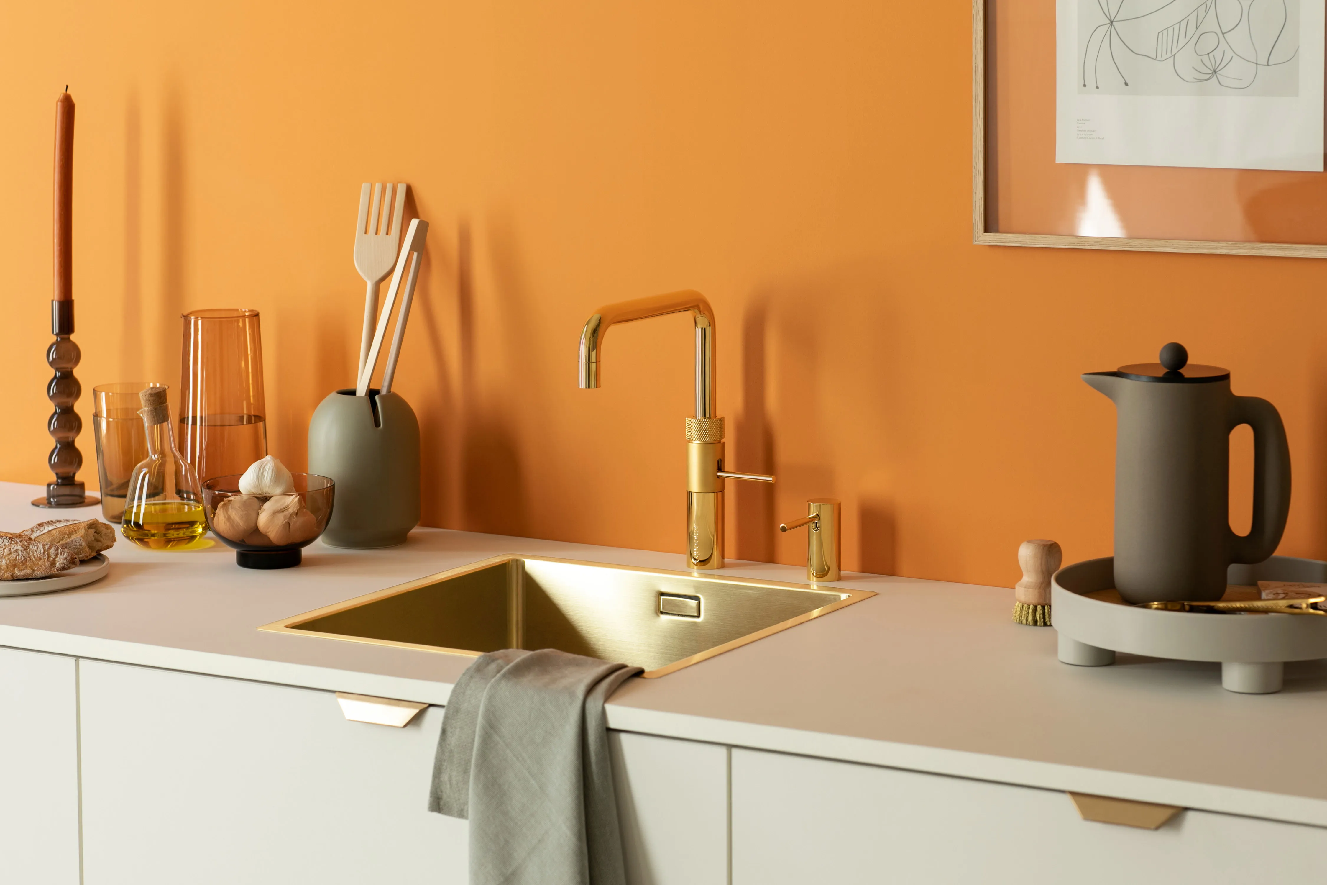 Goldener Quooker-Wasserhahn mit passendem Spülbecken und Seifenspender vor einer warmen, orangefarbenen Wand